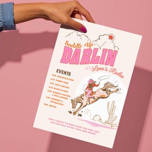 Könnte beinhalten: Ein rosa-cremefarbenes Rodeo-Event-Poster mit dem Text "Saddle Up Darlin" in Pink. Das Poster enthält eine Grafik eines Rodeo-Reiters und eine Liste von Veranstaltungen. Das Poster wird von einer Hand mit rosa Nagellack gehalten.