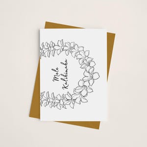 May include: A white card with a black and white floral wreath and the text "Mele Kalikimaka". The card is sitting on a gold envelope.