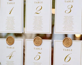 May include: A gold-framed mirror seating chart displaying six white cards labeled Table 1 through 6. Each card features gold text and a gold wax seal at the top. The mirror is positioned upright, reflecting soft ambient lights in the background.