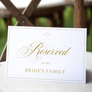 Op de afbeelding: Een witte kaart met gouden letters met de tekst "Reserved for the Bride's Family" wordt op een stoel weergegeven. De kaart heeft een gouden rand en het woord "Reserved" is in elegant schrift. De stoel is van hout en heeft een lichtgekleurd kussen.