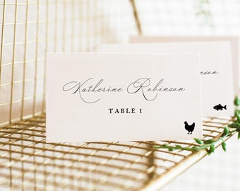 Op de afbeelding: Een wit rechthoekig naamkaartje met de in zwart gedrukte naam Katherine Robinson, de tekst TABLE 1 en een klein zwart silhouet van een kip. Een tweede identiek kaartje is erachter zichtbaar met een vissilhouet.