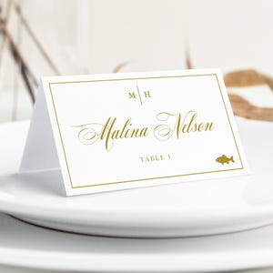 Op de afbeelding: Een witte gevouwen naamkaartje met een gouden rand, voorzien van de initialen M H, de naam Malina Nelson in gouden script, de tekst TABLE 1 en een klein gouden visicoontje. Het kaartje staat op een wit bord.