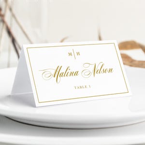 Op de afbeelding: Een witte gevouwen naamkaartje met een gouden rand, voorzien van de initialen M H, de naam Malina Nelson in gouden script en de tekst TABLE 1. Het kaartje staat op een wit bord, met de merknaam White Orchids Paperie zichtbaar.