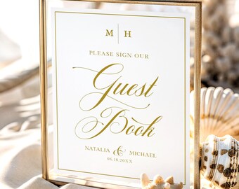 Op de afbeelding: Een rechthoekige gouden metalen lijst met een wit bordje met gouden tekst: M H, PLEASE SIGN OUR Guest Book, NATALIA & MICHAEL en 06.18.20XX. Het bordje heeft een dunne gouden rand en elegante cursieve letters voor de woorden Guest Book.