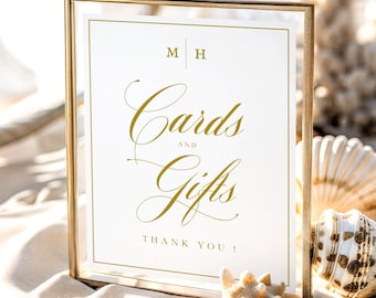 Op de afbeelding: Een rechthoekige goudkleurige metalen lijst bevat een wit bord met gouden letters. De tekst luidt M H, Cards and Gifts en THANK YOU!. Het bord is omringd door diverse schelpen op een lichte ondergrond, wat een strandthema creëert.