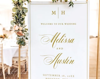 Op de afbeelding: Een wit welkomstbord voor een bruiloft met gouden letters en een gouden rand, opgehangen aan een goudkleurig metalen frame. Het bord bevat de initialen M H, de tekst Welcome to our wedding, de namen Melissa en Austin en de datum September 28, 20XX.