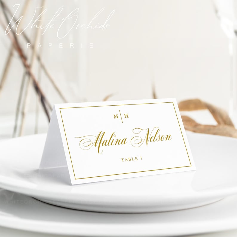 Op de afbeelding: Een witte gevouwen naamkaartje met een gouden rand, voorzien van de initialen M H, de naam Malina Nelson in gouden script en de tekst TABLE 1. Het kaartje staat op een wit bord, met de merknaam White Orchids Paperie zichtbaar.