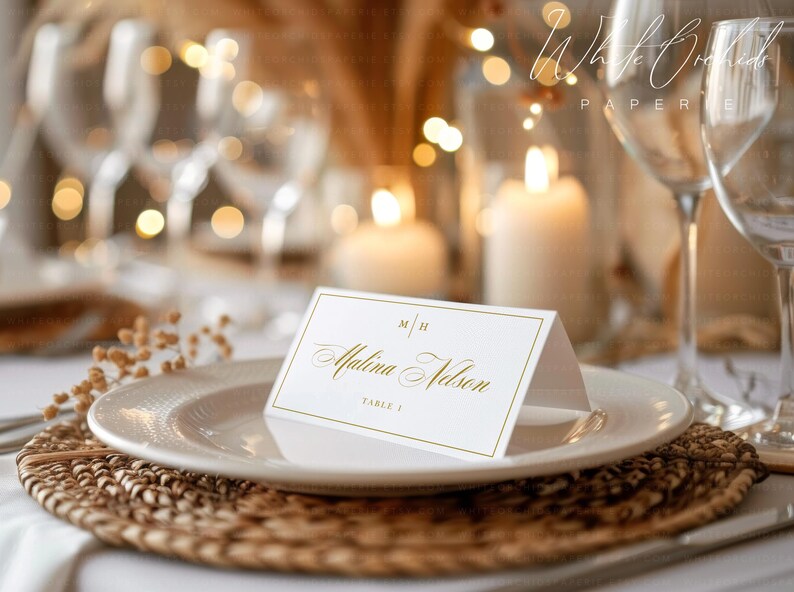 Op de afbeelding: Een witte gevouwen naamkaartje met goudfolie belettering met de naam Malina Nelson, de initialen M H en de tekst TABLE 2. Het kaartje staat in het midden op een wit bord dat op een rond gevlochten placemat rust met een wazige achtergrond.