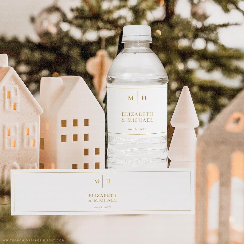May include: A clear water bottle with a white label featuring gold text that reads "M H" above "ELIZABETH & MICHAEL" and a date. The image also shows white decorative houses and a small white tree.