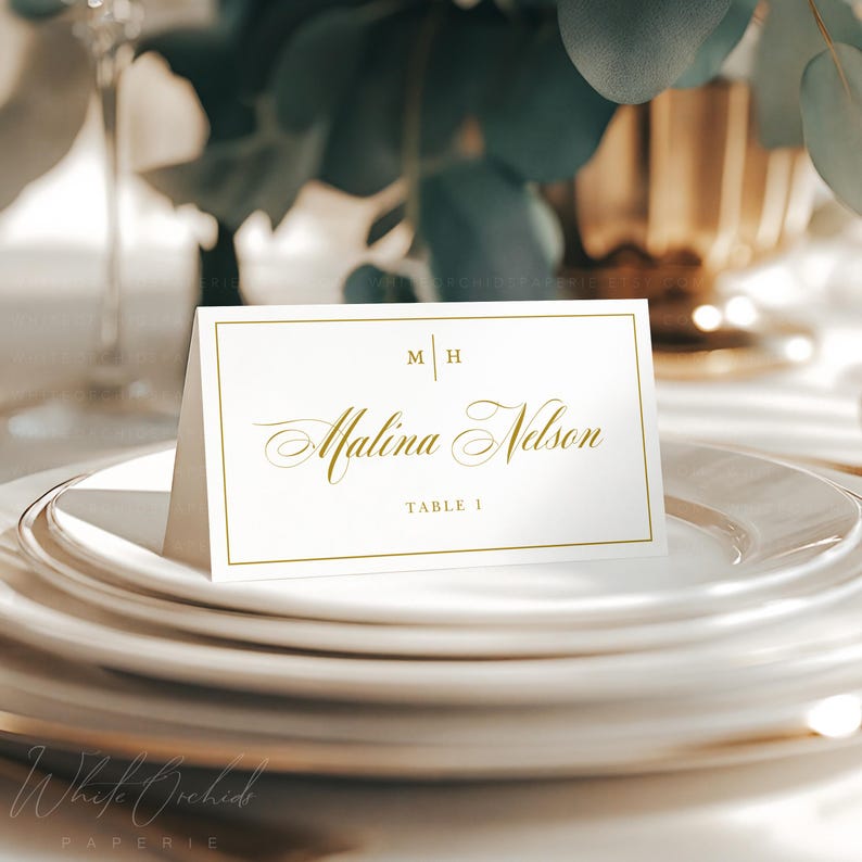 Op de afbeelding: Een witte rechthoekige gevouwen naamkaartje met een gouden rand. De kaart bevat de gouden tekst M | H bovenaan, de naam Malina Nelson in een gouden scriptlettertype in het midden en de tekst TABLE 1 in goud onderaan.