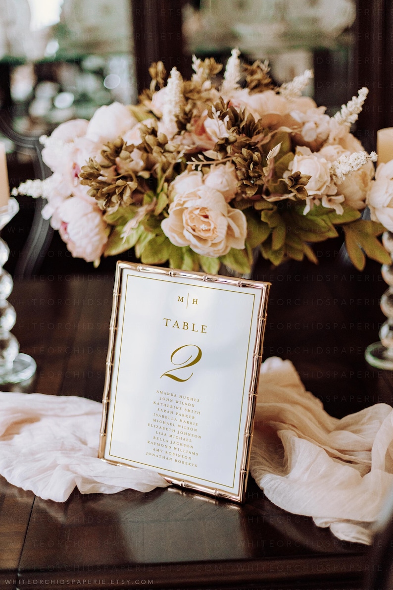 May include: A gold-framed table number card with the number 2, featuring a list of names. The card is set on a dark wooden table, with a soft, draped fabric and a floral arrangement in the background. The flowers are in shades of pink and cream.