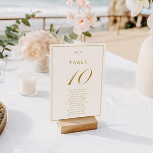 May include: A wedding table setting with a cream-colored table number card that reads "TABLE 10" in gold script. The card is propped on a wooden base. The table is decorated with flowers, candles, and a vase.