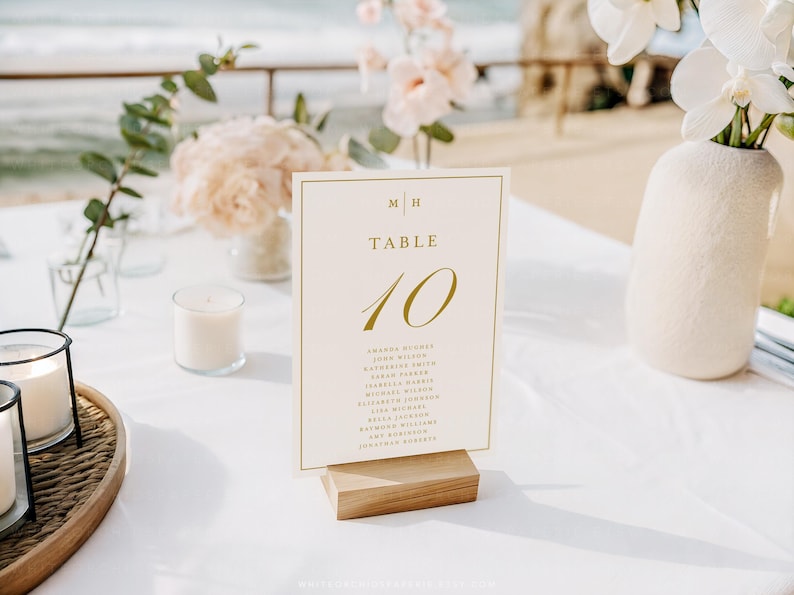 May include: A wedding table setting with a cream-colored table number card that reads "TABLE 10" in gold script. The card is propped on a wooden base. The table is decorated with flowers, candles, and a vase.