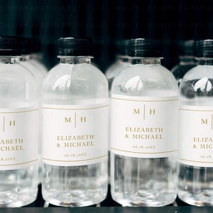 May include: Clear plastic water bottles with black caps. Each bottle has a white label with gold lettering that reads "M H" and "ELIZABETH & MICHAEL 06.18.20XX". The bottles are arranged in a row.