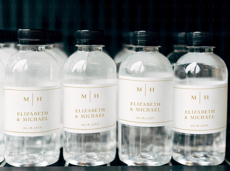 May include: Clear plastic water bottles with black caps. Each bottle has a white label with gold lettering that reads "M H" and "ELIZABETH & MICHAEL 06.18.20XX". The bottles are arranged in a row.