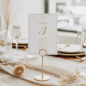 May include: A table number card with gold lettering and a gold border, held by a gold metal stand. The card reads "TABLE 3" and lists names. The background includes a table setting with glassware and floral arrangements.