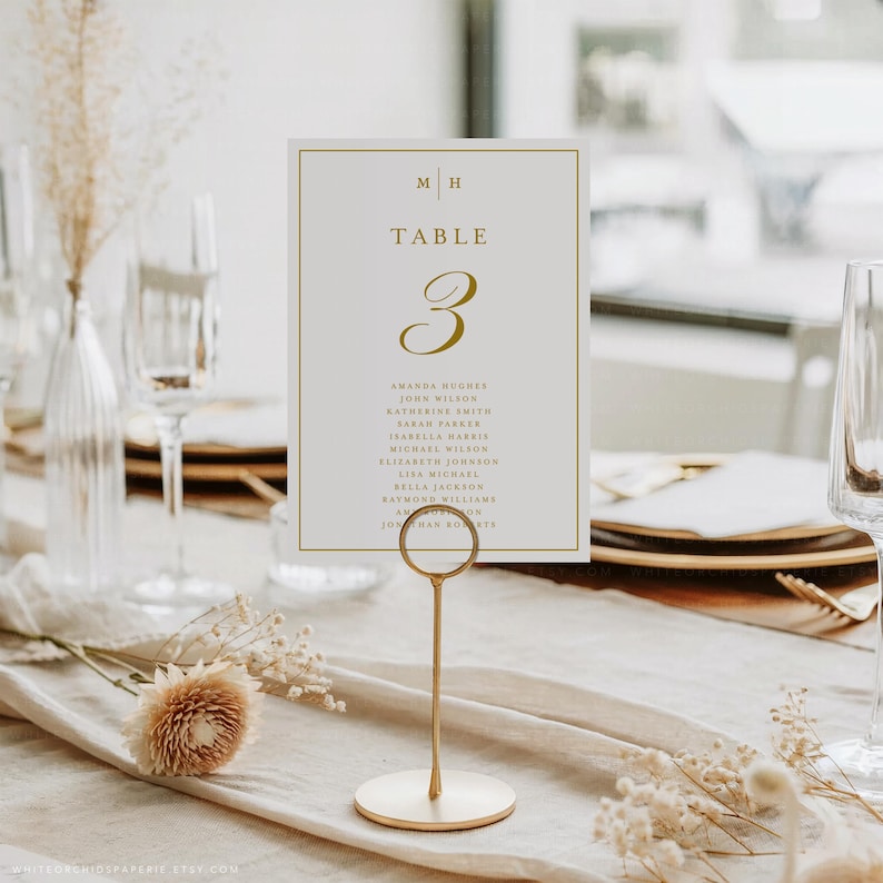 May include: A table number card with gold lettering and a gold border, held by a gold metal stand. The card reads "TABLE 3" and lists names. The background includes a table setting with glassware and floral arrangements.