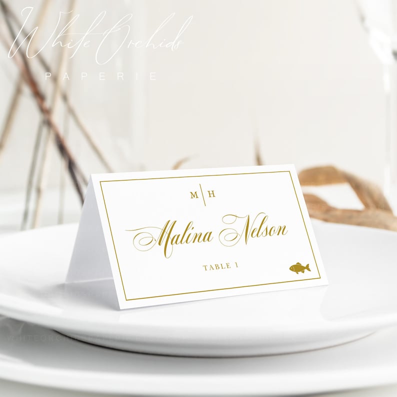 Op de afbeelding: Een witte gevouwen naamkaartje met een gouden rand, voorzien van de initialen M H, de naam Malina Nelson in gouden script, de tekst TABLE 1 en een klein gouden visicoontje. Het kaartje staat op een wit bord.