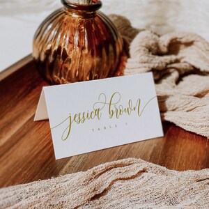 May include: A white place card with gold calligraphy that reads "jessica brown" and "table 1". The card sits on a wooden tray with a tan fabric and a brown glass vase with dried flowers. The Etsy shop name is visible.