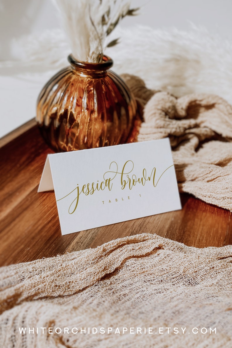 May include: A white place card with gold calligraphy that reads "jessica brown" and "table 1". The card sits on a wooden tray with a tan fabric and a brown glass vase with dried flowers. The Etsy shop name is visible.