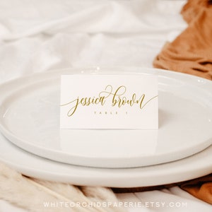 May include: A white place card with gold calligraphy that reads "jessica brown" and "table 1" is placed on a white plate. The plate is on a white surface with a brown cloth in the background. The Etsy shop name is at the bottom.
