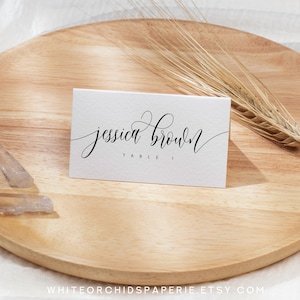 May include: A white place card with the name "Jessica Brown" in elegant black script, and "TABLE 1" below. The card rests on a wooden tray with a wheat stalk and clear crystals. The Etsy shop name is visible at the bottom.