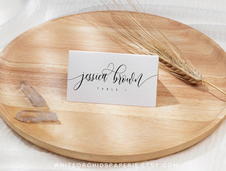 May include: A white place card with the name "Jessica Brown" in elegant black script, and "TABLE 1" below. The card rests on a wooden tray with a wheat stalk and clear crystals. The Etsy shop name is visible at the bottom.