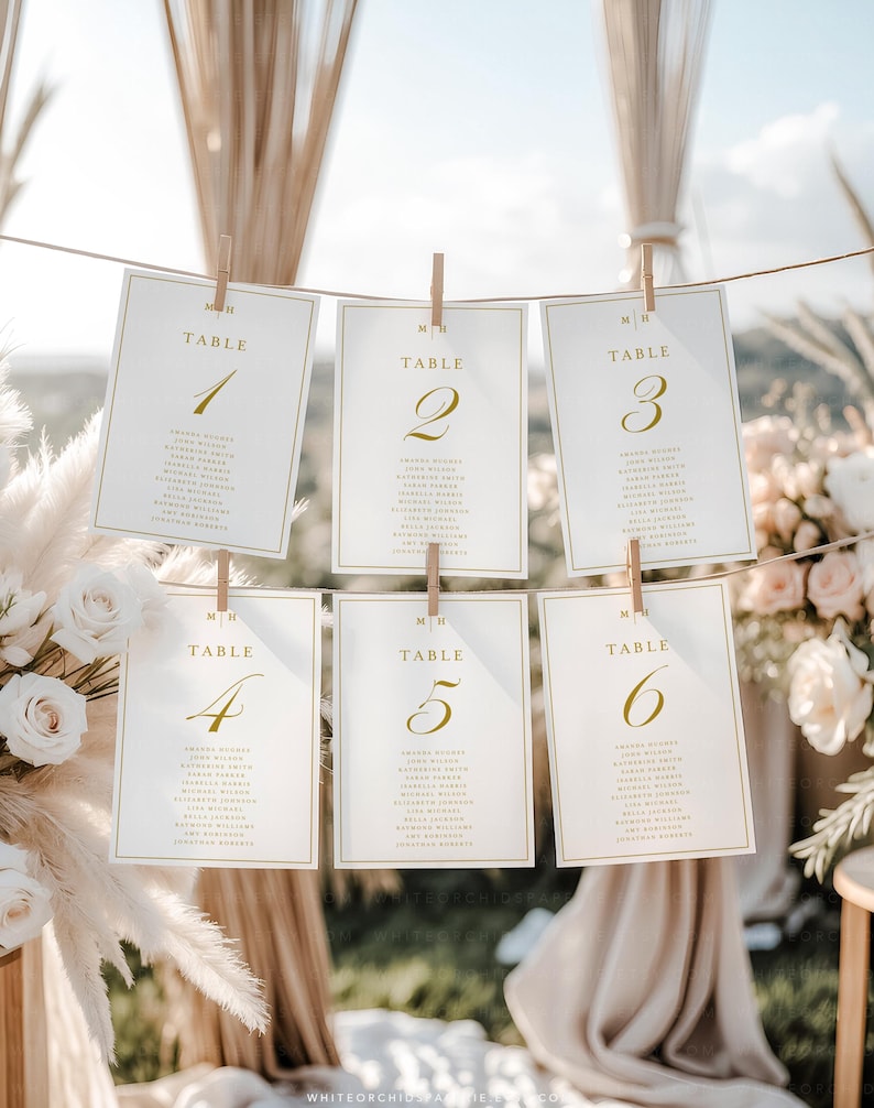 May include: Six white table cards with gold lettering and numbers 1-6 are clipped to a string with wooden clothespins. Each card lists names below the table number. The background includes floral arrangements and a blurred outdoor setting.