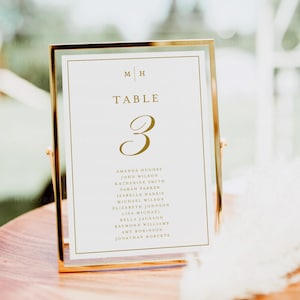May include: A gold-framed table number card with the number 3 in gold script. The card lists names under the table number. The top of the card has the initials "M H" and the word "TABLE". The card is displayed on a wooden surface.