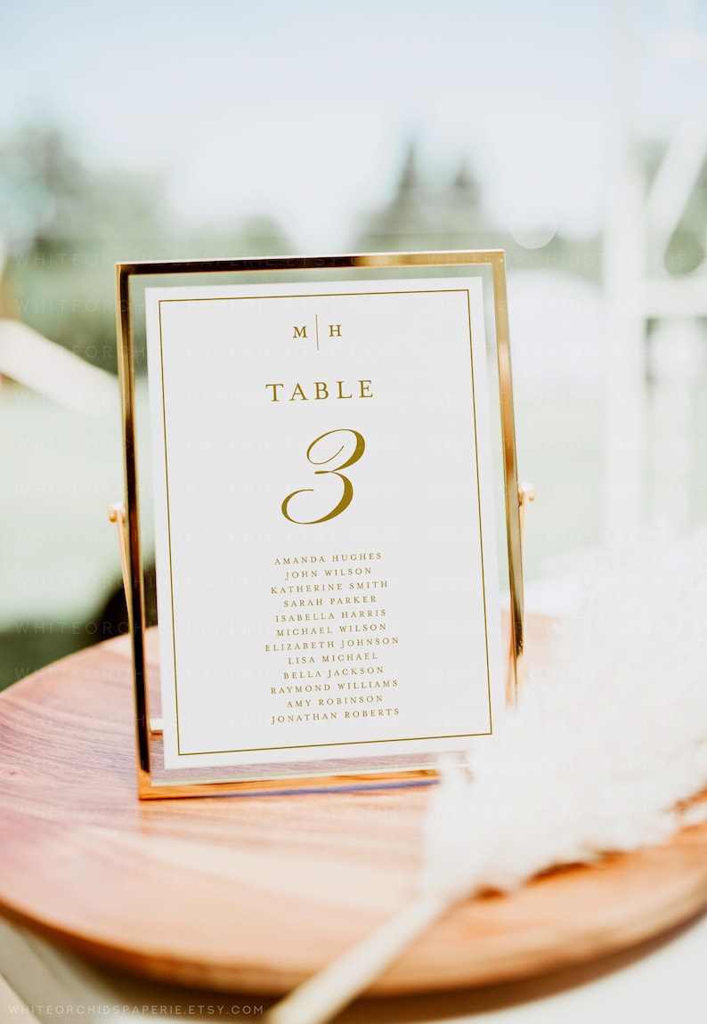 May include: A gold-framed table number card with the number 3 in gold script. The card lists names under the table number. The top of the card has the initials "M H" and the word "TABLE". The card is displayed on a wooden surface.
