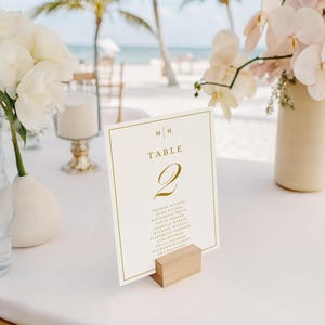 May include: A table setting with a white tablecloth, featuring floral arrangements and a table number card. The card, held by a wooden block, displays "Table 2" in gold script, with a list of names below. White and pink flowers are in vases.