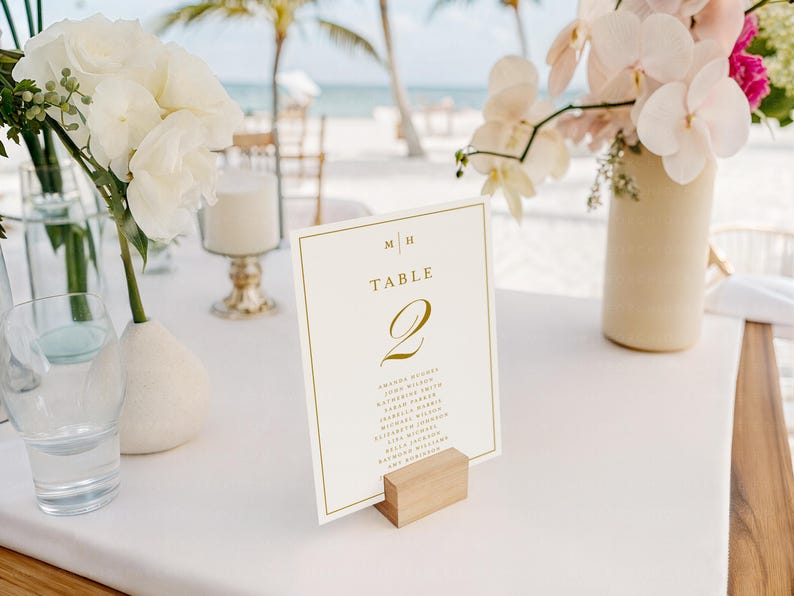 May include: A table setting with a white tablecloth, featuring floral arrangements and a table number card. The card, held by a wooden block, displays "Table 2" in gold script, with a list of names below. White and pink flowers are in vases.