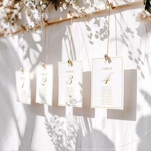 May include: Elegant wedding table number cards. Each card is white with gold lettering and a gold border. The cards are suspended by gold clips and string. The numbers 1, 2, 3, and 4 are visible, along with guest names.