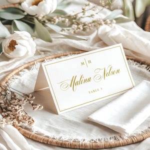 Op de afbeelding: Een witte gevouwen naamkaartje met een gouden rand, voorzien van de initialen M H, de naam Malina Nelson in gouden script en de tekst TABLE 1. Het kaartje staat op een geweven ronde schaal met witte stof en witte bloemen.