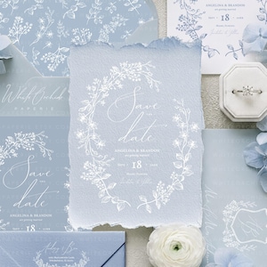May include: A light blue Save the Date card with deckled edges featuring white floral line art, elegant script text, and the names Angelina and Brandon. The stationery set includes matching envelopes and a white ring box with a diamond ring.