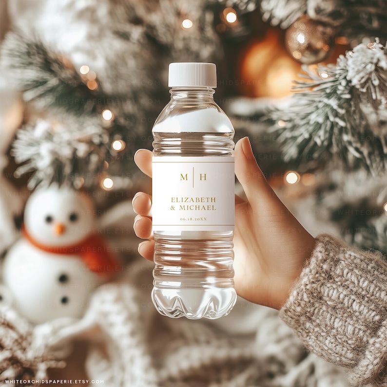 May include: A clear plastic water bottle with a white cap and a label featuring the initials "M H" and the names "ELIZABETH & MICHAEL" in gold lettering. The bottle is held in a hand, with a blurred background of a snowman and Christmas decorations.