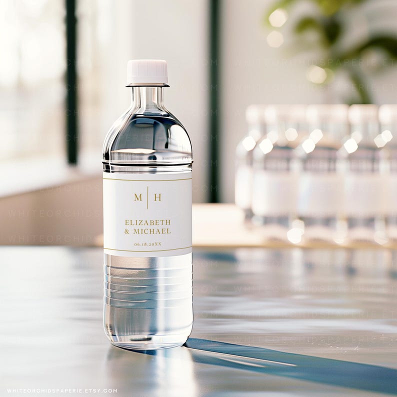 May include: A clear plastic water bottle with a white cap and a label featuring the initials "M H" and the names "Elizabeth & Michael" in gold lettering. The date "06.18.20XX" is also printed on the label. Several similar bottles are visible in the background.