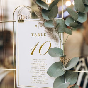 May include: A wedding table number 10 card with gold text and a gold border, held by a gold clip. The card includes a list of names. Eucalyptus and dried flowers decorate the card.