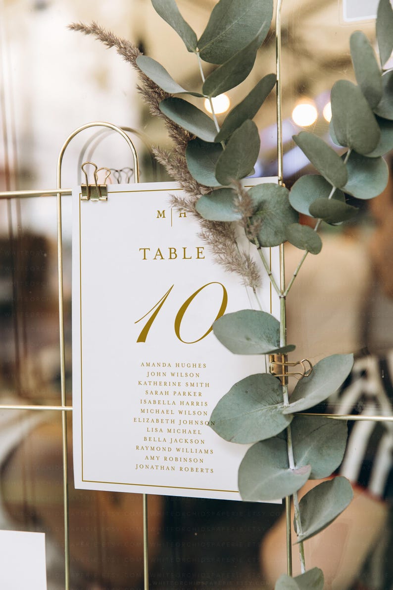 May include: A wedding table number 10 card with gold text and a gold border, held by a gold clip. The card includes a list of names. Eucalyptus and dried flowers decorate the card.