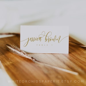 May include: A white place card with gold calligraphy that reads "jessica brown" and "table 1". The card is on a wooden surface with a wheat stalk. The Etsy shop name is visible at the bottom.