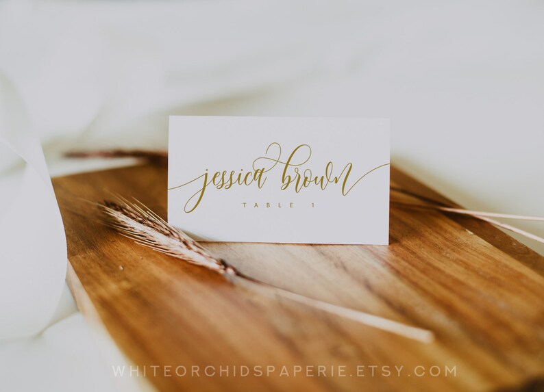 May include: A white place card with gold calligraphy that reads "jessica brown" and "table 1". The card is on a wooden surface with a wheat stalk. The Etsy shop name is visible at the bottom.