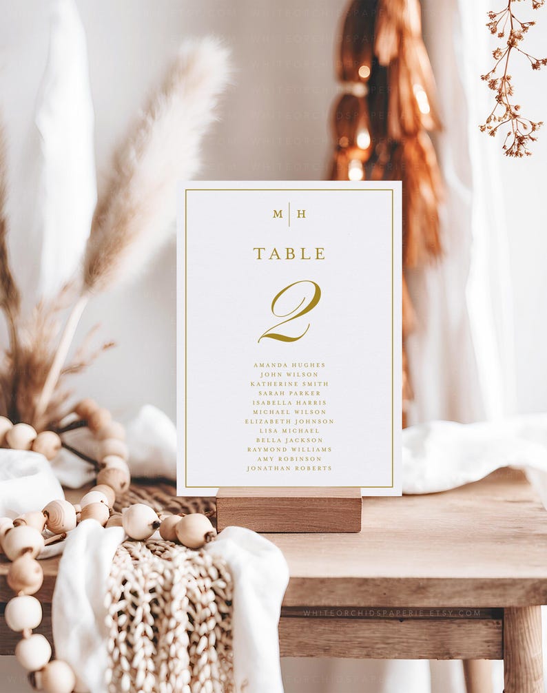 May include: A table number card with gold lettering and a gold border. The card displays the number "2" and the word "TABLE". The names of guests are listed below the number. The card is displayed on a wooden block.