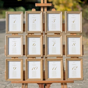 May include: Twelve framed table number signs displayed on a wooden easel. Each gold-framed sign has a white background with a table number and guest names in gold script. The numbers range from 1 to 12, suitable for a wedding or event.