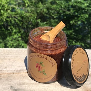May include: A glass jar with a brown label that reads "Schisandra Miel" and "Honey-based Botanical". The jar is filled with a brown, grainy substance and a wooden spoon is resting on top. The jar has a black lid with a brown label that reads "Warrior Peak Botanicals".