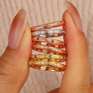 May include: A stack of six thin metal rings featuring a repeating marquise-shaped pattern with milgrain detailing. The rings are crafted in various gold tones, including yellow, rose, and white, and are accented with small embedded gemstones.
