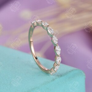 May include: A delicate, rose gold band with a row of marquise-shaped diamonds set in a prong setting. The band is on a light blue surface.
