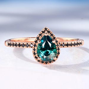 May include: Rose gold ring featuring a pear-shaped teal gemstone, surrounded by a halo of black stones. The band is also adorned with small black stones, creating a delicate and elegant design. Fine jewelry.
