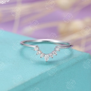 May include: A delicate silver ring with a curved row of small, clear diamonds. The ring is on a light blue surface.