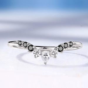 May include: A curved silver band ring featuring a central pear-cut white stone flanked by two round white stones, with three smaller round black stones set along each side of the band, creating a symmetrical V-shaped design on a white surface.