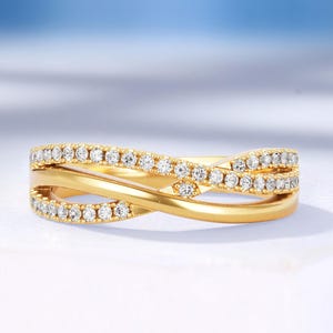 May include: A gold-colored ring with a crisscross design. The ring features a row of small, clear gemstones set along the top and bottom bands. The ring is displayed on a white surface with a soft blue background.